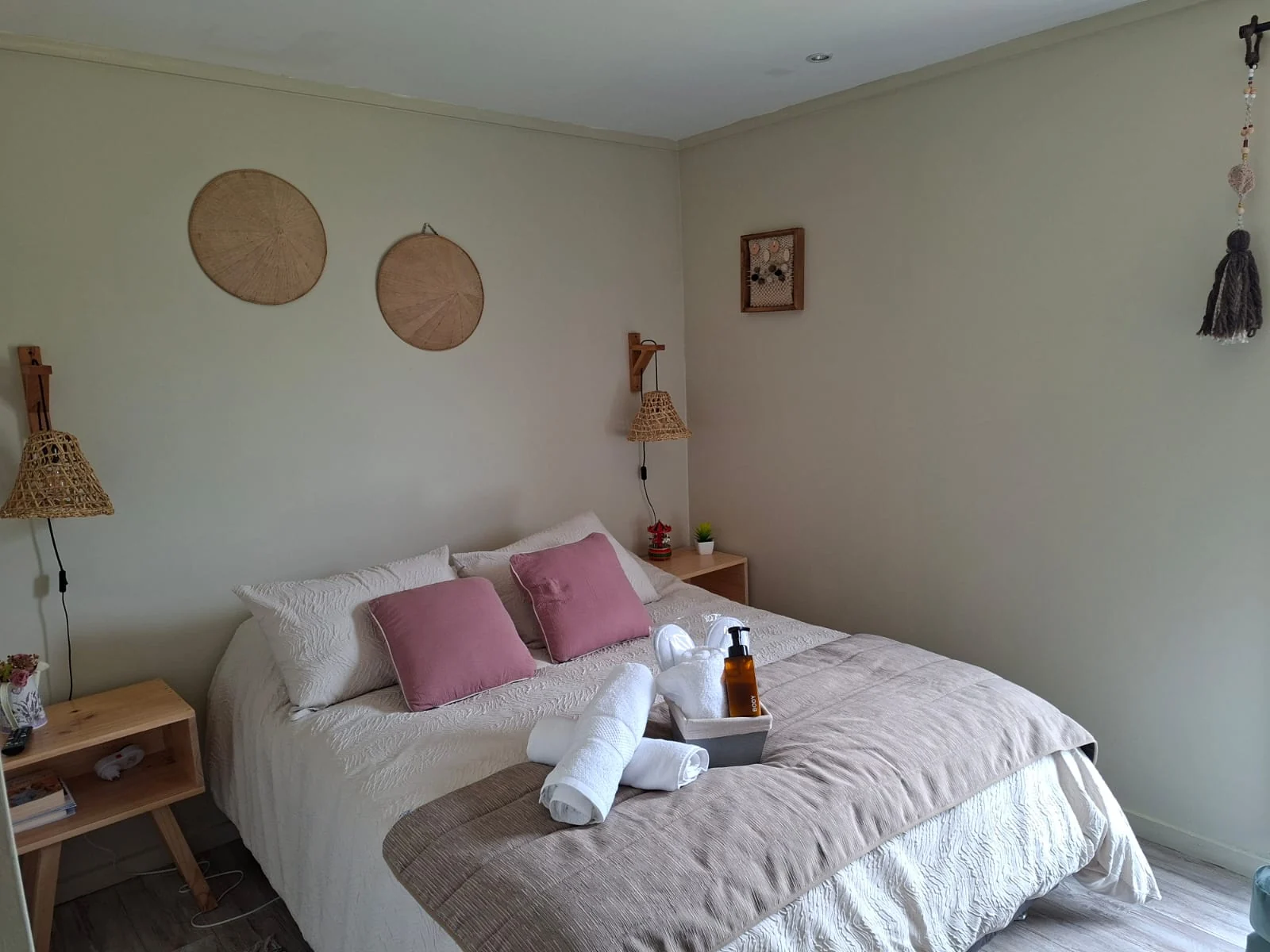 Cama matrimonial con cojines rosados y toallas, con sombreros de mimbre en la pared