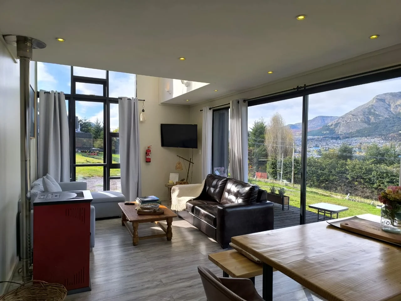 Living con estufa roja, sillón de cuero y comedor junto a ventanales de doble altura con vista a las montañas